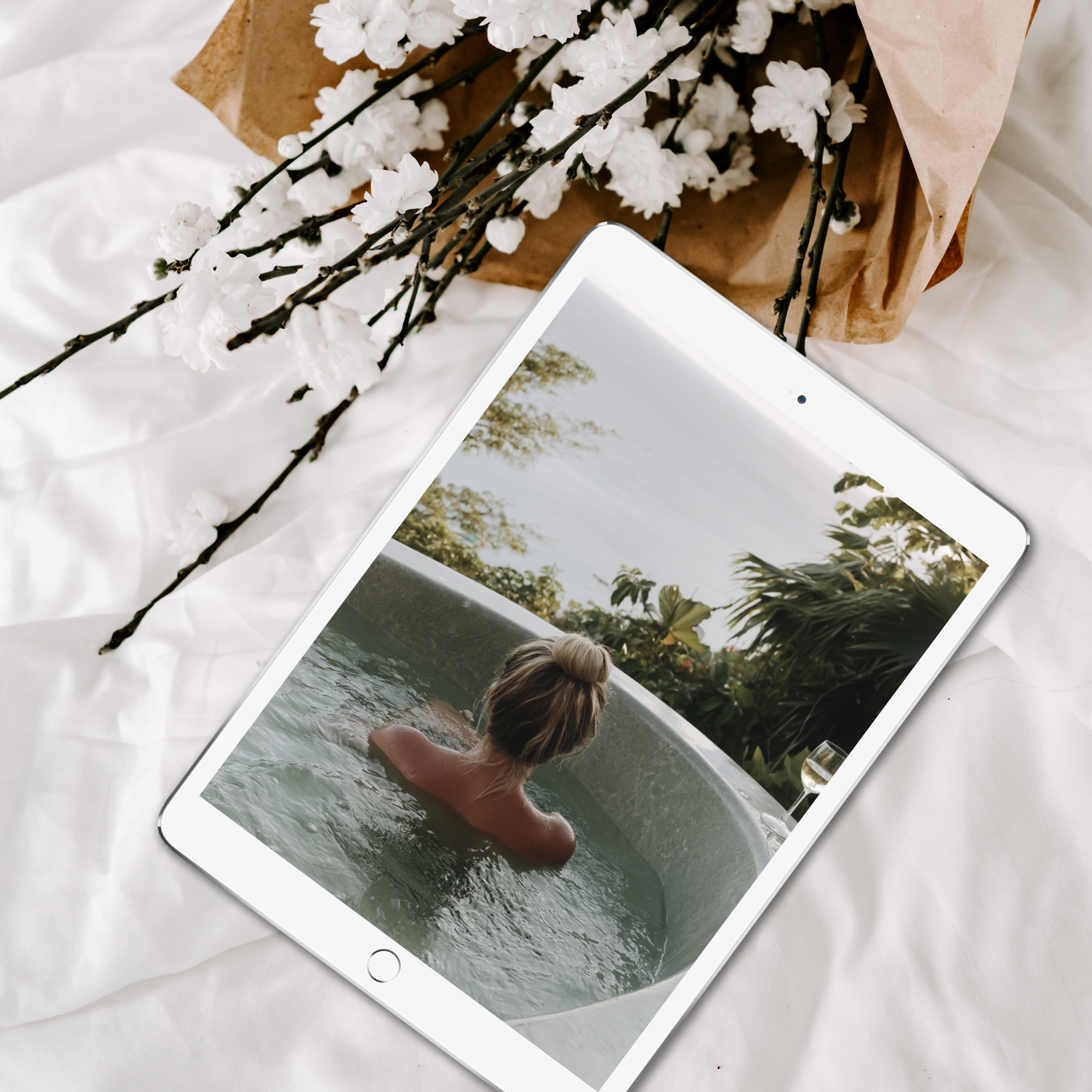 A tablet displaying a photo of a woman in a hot tub is placed on a white bedspread next to a bouquet of white flowers.